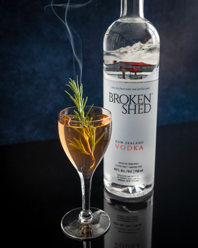 An orange coloured cocktail with a smoking rosemary sprig as garnish, sitting next to a bottle of Broken Shed Vodka